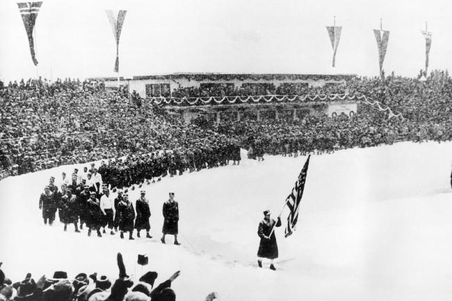 Олимпийские игры 1936, Гармиш-Партенкирхен. Члены олимпийской сборной США маршируют за американским флагом на заснеженном лыжном стадионе во время церемонии открытия IV зимних Олимпийских игр в Гармиш-Партенкирхене в Германии 6 февраля 1936 года. В играх приняли участие 668 спортсменов из 28 стран.